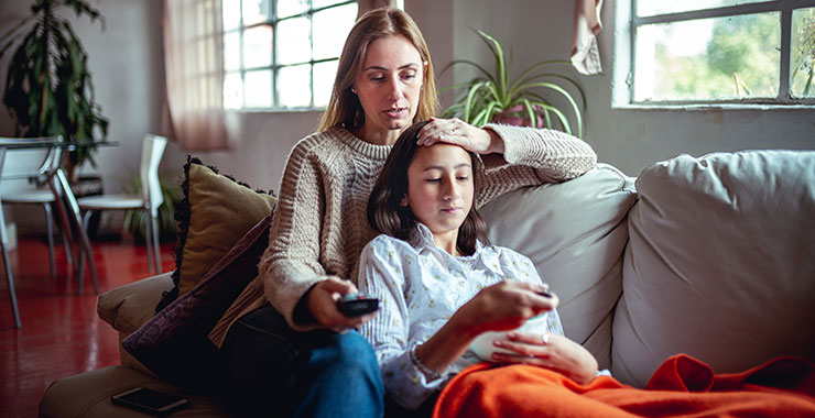 Daughter relaxes against mom on couch; both looking at daughter's phone screen.