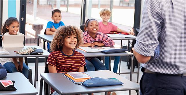 Elementary school students seated at desks in classroom, looking at teacher while smiling.