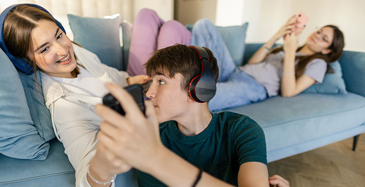 Three teenagers relaxing on couch; two are sharing a smart phone, smiling at screen.