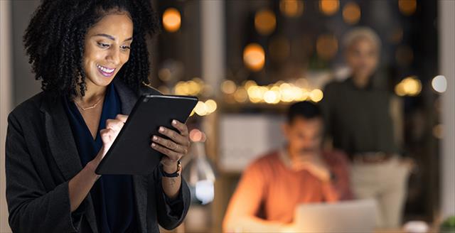 Woman reads a tablet at night with a smile on her face while her colleagues, who are out of focus, work in the background