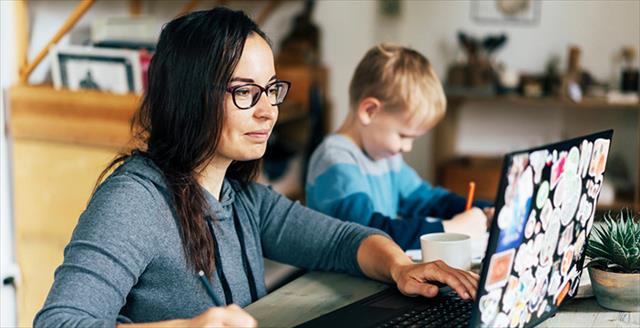 Woman working online off a laptop and taking notes with a child sitting next to her coloring