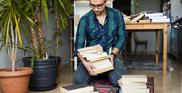 Young man kneeling on the floor of his cozy home office organizing stacks of large books, his office plants are thriving