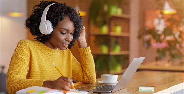 Young person studying on a laptop with headphones on taking notes with a nuanced side-smile