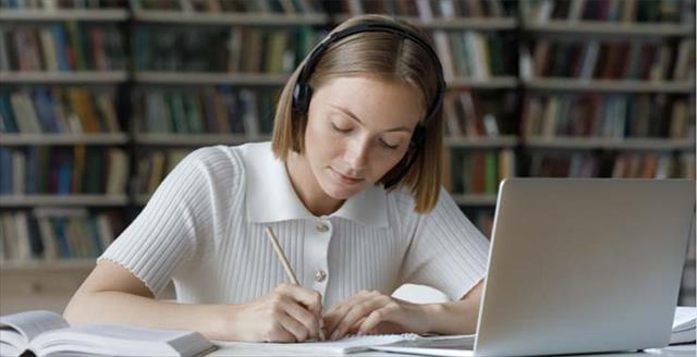 Young student works on a laptop with headphones on, sitting in a library with determined focus