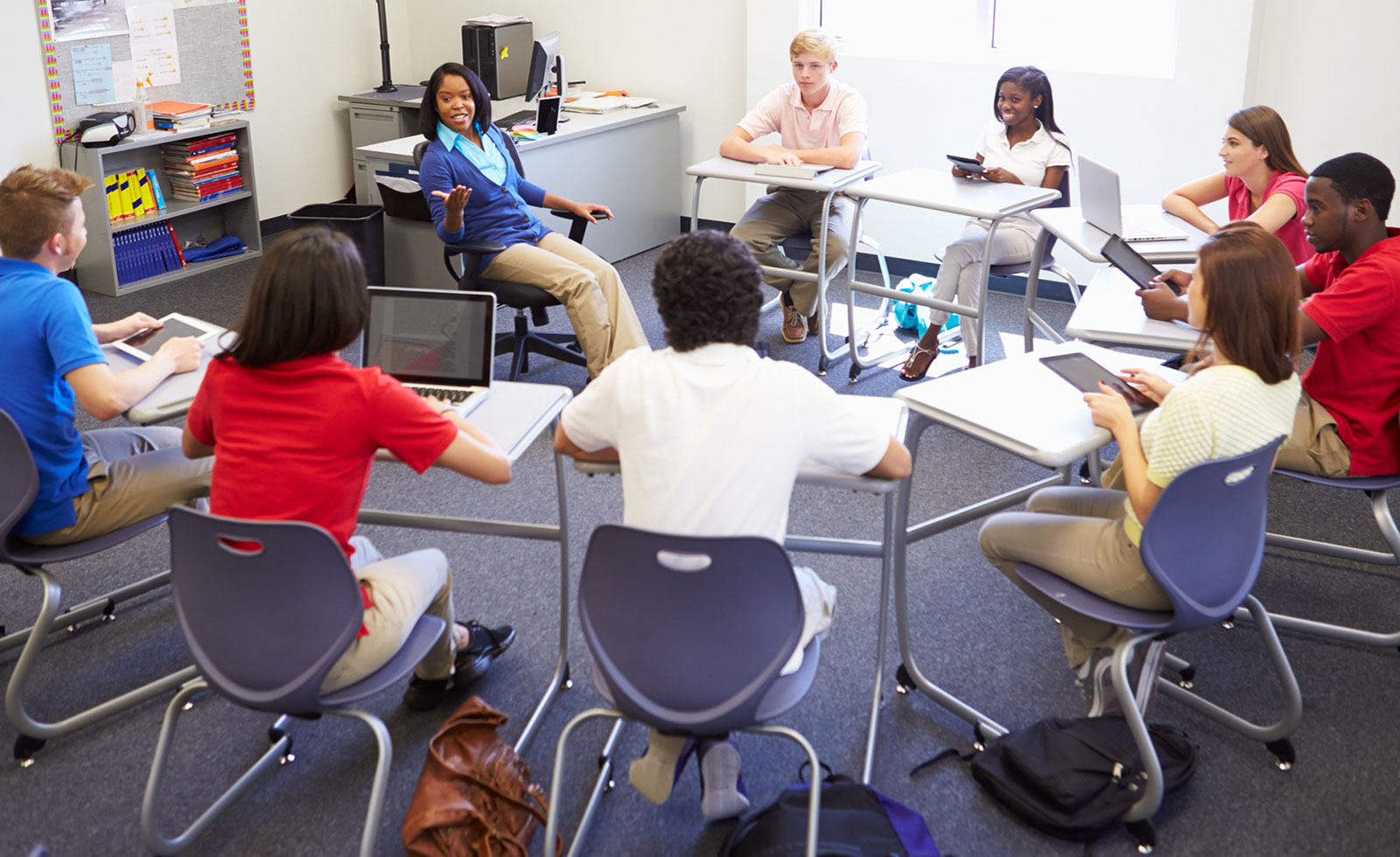 Teacher seated in chair talking to students in classroom.