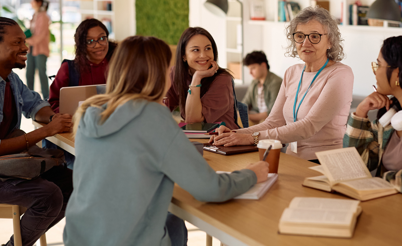 Older professor sitting at wooden table surrounded by diverse group of college students.